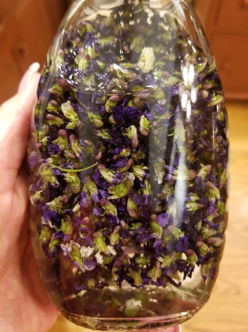 Puede incluir: Una botella de vidrio llena de p&eacute;talos de flores moradas y verdes sumergidos en un l&iacute;quido transparente.
