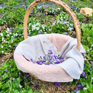Puede incluir: Una cesta de mimbre llena de una toalla rosa suave y una colecci&oacute;n de flores silvestres moradas y blancas. La cesta est&aacute; sentada sobre un lecho de hierba verde y flores silvestres.