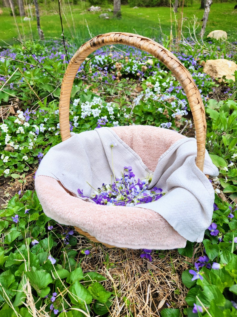 Puede incluir: Una cesta de mimbre llena de una toalla rosa suave y una colecci&oacute;n de flores silvestres moradas y blancas. La cesta est&aacute; sentada sobre un lecho de hierba verde y flores silvestres.
