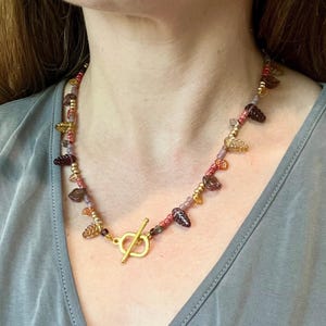 May include: A necklace with a gold toggle clasp. The necklace is adorned with small, round beads in pink, purple, and gold, alongside leaf and teardrop-shaped beads. The necklace is worn around the neck.