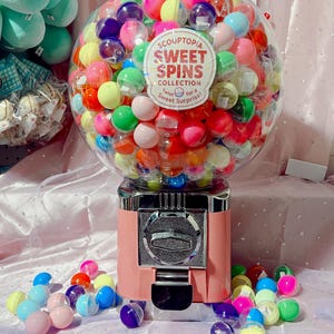 Puede incluir: Una máquina de chicles rosa llena de cápsulas de plástico de colores. La máquina tiene una ranura para monedas plateada y una pegatina "SCOUPTOPIA SWEET SPINS COLLECTION". Varias cápsulas y monedas están esparcidas alrededor de la base de la máquina.