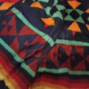 Può includere: Primo piano di una morbida coperta fantasia. La coperta presenta uno sfondo blu navy con forme geometriche rosse, arancioni e verdi. I bordi hanno un motivo a righe nelle tonalità del rosso, dell'arancione e del giallo.