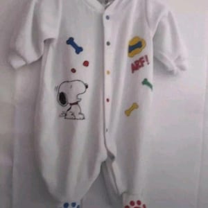 May include: White baby onesie featuring a Snoopy graphic, colorful bone and dot accents, and the word "ARF!". The footed pajamas have blue and red paw prints on the feet. The onesie has a button-up front.