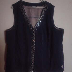 May include: A dark blue denim vest featuring a V-neckline and a front button closure. The collar, front placket, and hem are embellished with silver-toned rhinestones. A small silver heart-shaped emblem is visible on the lower right side.