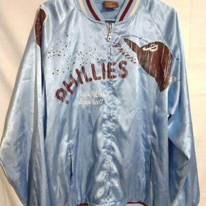 Peut inclure: Veste de baseball en satin bleu clair avec le mot "Phillies" en lettres marron. La veste présente un col, des poignets et une taille rayés marron et blancs. Un logo d'équipe de baseball est sur le côté droit.