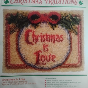 May include: A WonderArt latch hook wall hanging kit titled Christmas Traditions. The package features a finished rug design with the text Christmas is Love in red, surrounded by green holly leaves and red bows on a cream background, 27 by 20 inches.