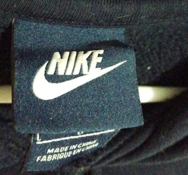 May include: A navy blue tag featuring the white Nike logo and the word "NIKE" in white block letters. Below the logo, the tag reads "MADE IN CHINA" and "FABRIQUE EN CHINE". The tag is attached to a dark blue garment.