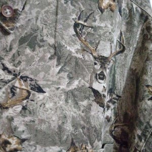 May include: Camouflage button-up shirt with a deer head design. The shirt features a pattern of green, brown, and grey, creating a woodland camouflage effect. The deer head is prominently displayed on the fabric.