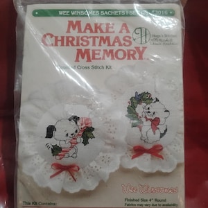 May include: A packaged cross-stitch kit featuring two round designs with Christmas themes. One design shows a puppy with a candy cane, the other a cat with a wreath. The kit includes instructions, white Aida cloth, eyelet, ribbon, felt, potpourri, needle, and DMC floss.