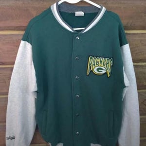 Puede incluir: Chaqueta verde y gris con botones y el logotipo de los Packers en la parte delantera. La chaqueta tiene mangas grises y un cuerpo verde. El cuello y los puños tienen rayas blancas y grises. La chaqueta está colgada en una percha blanca.