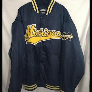 May include: Navy blue satin jacket with yellow accents. The word "Michigan" is prominently displayed across the chest in yellow script, with a matching "M" logo on the sleeve. The collar, cuffs, and hem feature yellow stripes.
