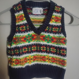 Può includere: Un gilet lavorato a maglia blu navy con scollo a V e un motivo geometrico colorato in rosso, giallo, verde e bianco. Il gilet è appeso a una gruccia grigia. L'etichetta recita "Kidstring".