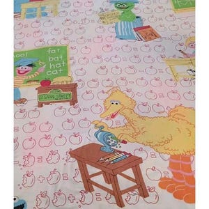 Op de afbeelding: Een wit stoffen laken met een herhalend patroon van rood omrande appels en Sesame Street-personages zoals Big Bird, Cookie Monster en Oscar the Grouch in schoolomgevingen met bureaus, boeken en schoolborden met de tekst fat bat hat cat.