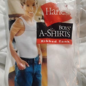 以下が含まれることがあります： Hanes Boys' A-Shirts リブタンクトップのパッケージ。 パッケージは透明なプラスチックで、シャツは白です。 パッケージには、白いタンクトップとブルージーンズを着た人の写真が掲載されています。