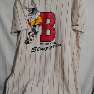 May include: Cream-colored baseball jersey with red pinstripes. Features a large red "B" and a Bugs Bunny graphic. The text "BURBANK Sluggers" is printed below the "B".