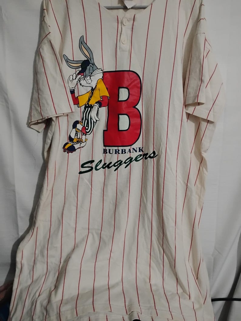 May include: Cream-colored baseball jersey with red pinstripes. Features a large red "B" and a Bugs Bunny graphic. The text "BURBANK Sluggers" is printed below the "B".