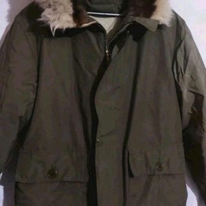 Puede incluir: Parka de invierno verde oliva con capucha ribeteada de piel. La chaqueta presenta cierre de cremallera, bolsillos abotonados y un diseño cálido y aislado. Una prenda de abrigo clásica para el frío.