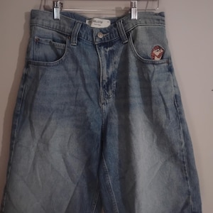 May include: Light blue denim shorts with a medium wash, featuring a button and zipper fly closure, five pockets, and a small embroidered graphic of a cartoon character on the front right pocket area. The inner label reads Hollister.