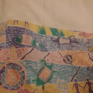 Puede incluir: Una sábana de tela blanca con un borde estampado colorido que muestra ilustraciones de una tortuga verde, un pez, un lagarto y figuras humanas. El patrón incluye formas geométricas, líneas azules onduladas y secciones amarillas.