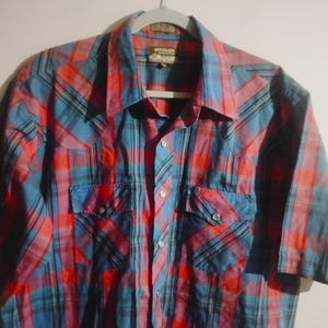 May include: A short-sleeved plaid shirt in shades of red, blue, and purple, hanging on a hanger. The shirt features two chest pockets with button closures and a classic collar. The plaid pattern is a mix of vertical and horizontal lines.