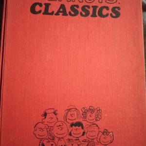 以下が含まれることがあります： 黒い文字で「PEANUTS CLASSICS」と書かれた赤い本。表紙には、チャーリー・ブラウンやスヌーピーなど、ピーナッツのキャラクターの白黒イラストが描かれています。