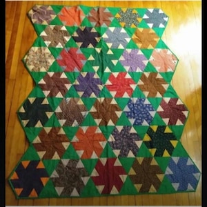 Puede incluir: Un edredón hecho a mano con un diseño de estrella. El edredón presenta un fondo verde con bloques en forma de estrella en varios colores, incluyendo marrón, rojo, naranja y azul. El edredón está colocado sobre un suelo de madera.