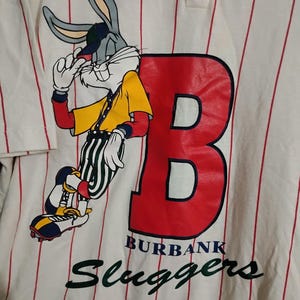 May include: White and red striped baseball jersey featuring Bugs Bunny in a yellow shirt and striped pants. The jersey has a large red "B" and the words "BURBANK Sluggers" in green.