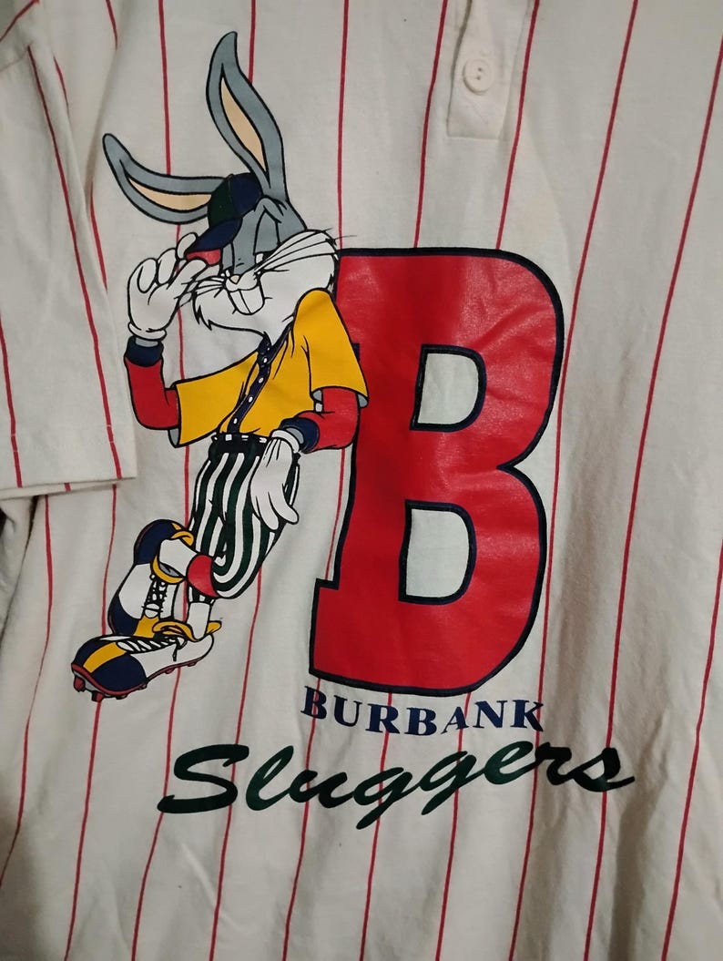 May include: White and red striped baseball jersey featuring Bugs Bunny in a yellow shirt and striped pants. The jersey has a large red "B" and the words "BURBANK Sluggers" in green.