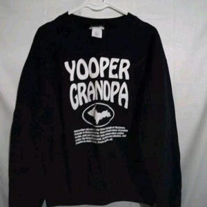 Puede incluir: Sudadera negra con el texto blanco "YOOPER GRANDPA". Debajo, un óvalo blanco con un gráfico de la Península Superior de Michigan. La sudadera está colgada en una percha blanca.