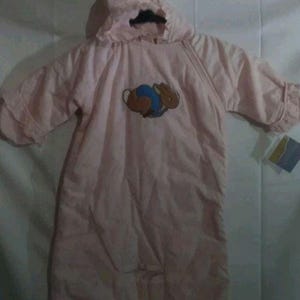 May include: A light pink baby bunting with a hood and short sleeves. The bunting features an embroidered design of a rabbit and a ball. The garment is hanging on a black hanger.