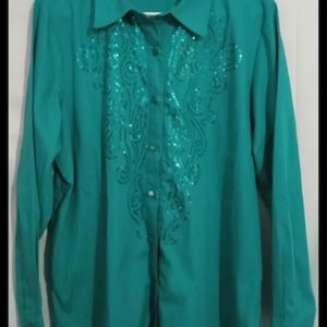 Puede incluir: Camisa abotonada de color turquesa con mangas largas. La camisa presenta un diseño de lentejuelas en la parte delantera y un cuello. La camisa está colgada en una percha blanca.