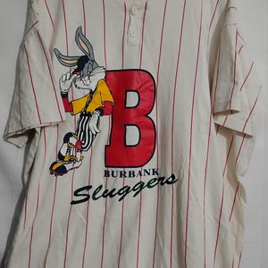 May include: Cream-colored baseball jersey with red pinstripes. Features a large red "B" with "BURBANK Sluggers" text and a Bugs Bunny graphic. The jersey has short sleeves and a button-up front.