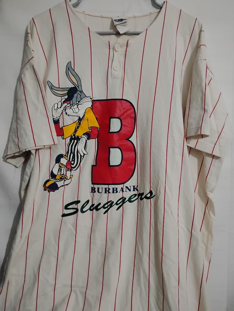 May include: Cream-colored baseball jersey with red pinstripes. Features a large red "B" with "BURBANK Sluggers" text and a Bugs Bunny graphic. The jersey has short sleeves and a button-up front.