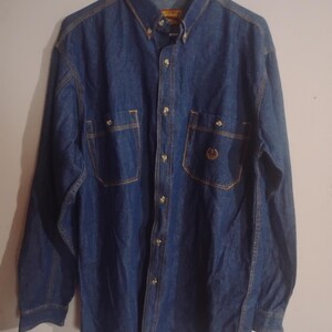 May include: A blue denim shirt with long sleeves, two chest pockets, and gold-colored buttons. The shirt has a button-down collar and a small logo on one pocket. The stitching is a contrasting gold color.