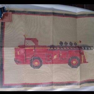 May include: A needlepoint canvas featuring a red fire truck design. The fire truck has black and yellow details, with a ladder and four lights on top. The background is beige with a red and black border.