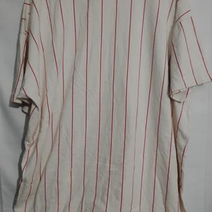 May include: Cream-colored baseball jersey with vertical red stripes. The jersey has short sleeves and a crew neck. The fabric appears to be a lightweight material, suitable for warm weather.