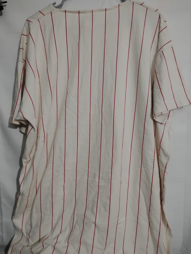 May include: Cream-colored baseball jersey with vertical red stripes. The jersey has short sleeves and a crew neck. The fabric appears to be a lightweight material, suitable for warm weather.