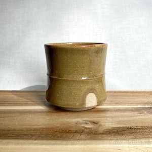 May include: A cylindrical ceramic cup with a glossy olive-green glaze. The vessel features a distinct horizontal ridge around the middle and a small, unglazed semi-circular arch detail at the base, revealing the light-coloured clay underneath.