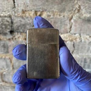 May include: A silver metal lighter with a brushed finish. The rectangular lighter is held by a hand wearing a blue glove. The background is a grey brick wall.