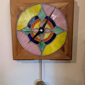 Può includere: Un orologio da parete quadrato in legno con un quadrante in vetro colorato. Il vetro presenta un design circolare con segmenti rosa, gialli, blu, verdi e arancioni. L'orologio ha lancette nere e un pendolo.