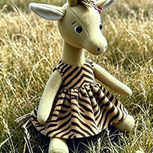Könnte beinhalten: Ein Giraffen-Stofftier mit beigem Körper, dunkelbraunen Augen und einer kleinen rosa Schleife am Kopf. Die Giraffe trägt ein ärmelloses Kleid mit einem schwarz-beigen Zebramuster, einer gerafften Taille und einem ausgestellten Rock.