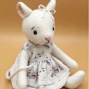 May include: A white felt mouse figurine with black eyes, a pink nose, and long whiskers. The mouse wears a white dress featuring a grey and pink floral pattern, accented with a white ribbon bow on its head and white lace trim at the hem.
