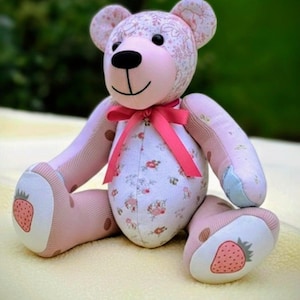 Peut inclure: Un ours en peluche fabriqué à partir de divers tissus, avec un motif floral rose sur la tête et le torse, des membres rayés roses et des coussinets blancs décorés de fraises. Un ruban rose vif est noué autour de son cou.