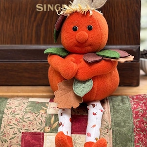 May include: A plush pumpkin doll with a round orange body, a beige stem, and black bead eyes. The doll features felt leaves in green and brown, a raffia accent, and long white legs patterned with small autumn leaves, ending in orange fabric feet.