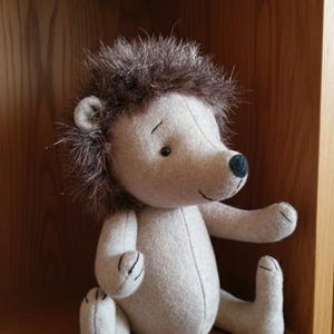 Op de afbeelding: Een pluche egelknuffel met een lichtbeige viltlichaam en donkerbruine imitatiebont stekels. Het speelgoed heeft een kleine zwart gestikte neus, eenvoudige zwarte ogen en gestikte details op de pootjes. Het zit in een zittende houding met de armen naar voren gestrekt.