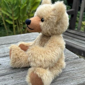 Puede incluir: Un oso de peluche con pelaje beige largo y desgreñado y un hocico de fieltro liso de color tostado. El oso presenta una nariz pequeña y redonda de color negro, una boca negra cosida y ojos oscuros y redondos. Está sentado de perfil con almohadillas de fieltro.