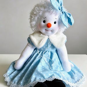 Puede incluir: Una muñeca de peluche de muñeco de nieve con cabeza blanca y difusa, nariz de zanahoria naranja brillante y ojos negros. La muñeca viste un vestido azul claro con lunares blancos, un cuello de piel sintética blanca, un lazo a juego y zapatos negros.