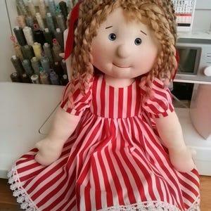 May include: A fabric doll with light skin, blue eyes, and curly brown hair styled with a large red ribbon. The doll wears a short-sleeved dress featuring vertical red and white stripes and a white lace trim along the bottom hem.