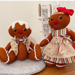 Könnte beinhalten: Zwei braune Lebkuchen-Stoffpuppen mit weißem Zuckerguss-Design. Die linke Puppe trägt eine gestreifte Fliege und Weste. Die rechte Puppe trägt eine rote Haarschleife und ein beiges Kleid mit Rentier-Aufdruck und Streifen. Ein Schild zeigt HAND MADE WITH.