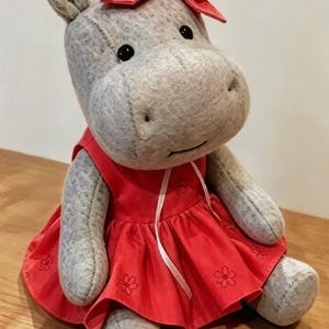 May include: A plush hippo toy made of light gray textured fabric with black bead eyes. The hippo wears a bright red sleeveless dress featuring a subtle embroidered flower pattern and a thin white ribbon tie, topped with a matching red bow.